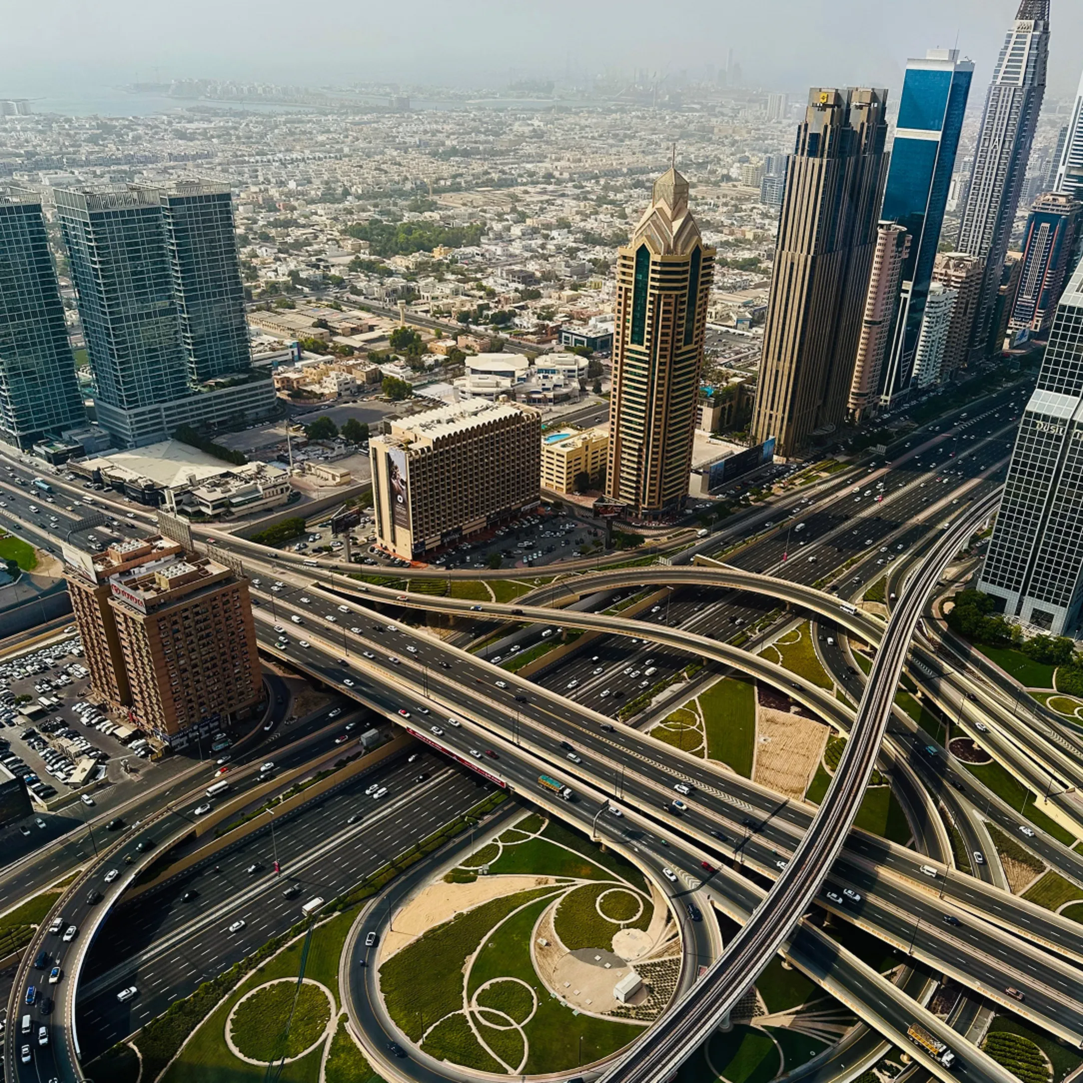 Aerial view of a complex highway interchange with multiple overpasses and circular green landscaping in an urban area with tall buildings and dense cityscape in the distance.