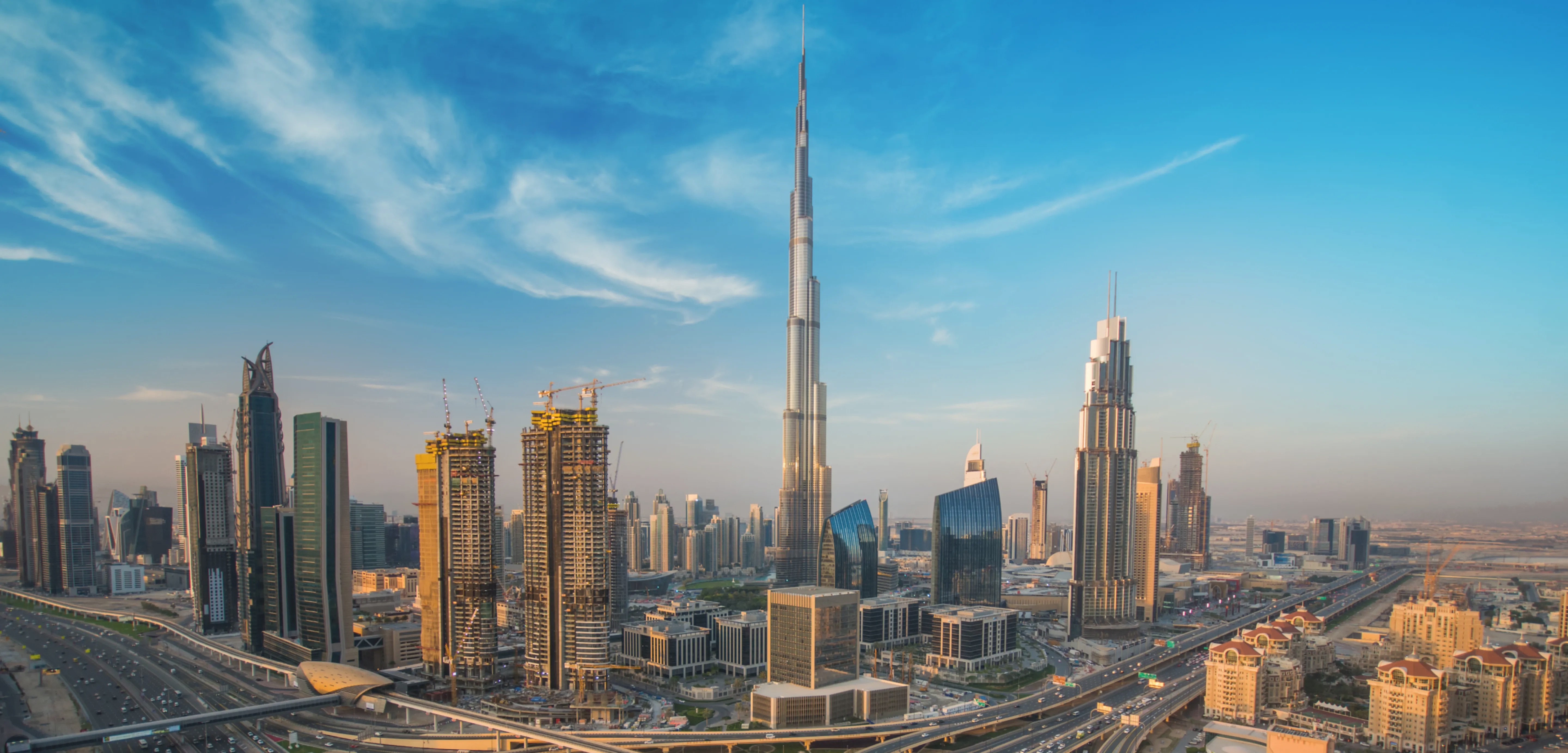Panoramic view of Dubai skyline featuring the Burj Khalifa, the tallest building, surrounded by modern skyscrapers, highways, and a partly cloudy blue sky at sunset.
