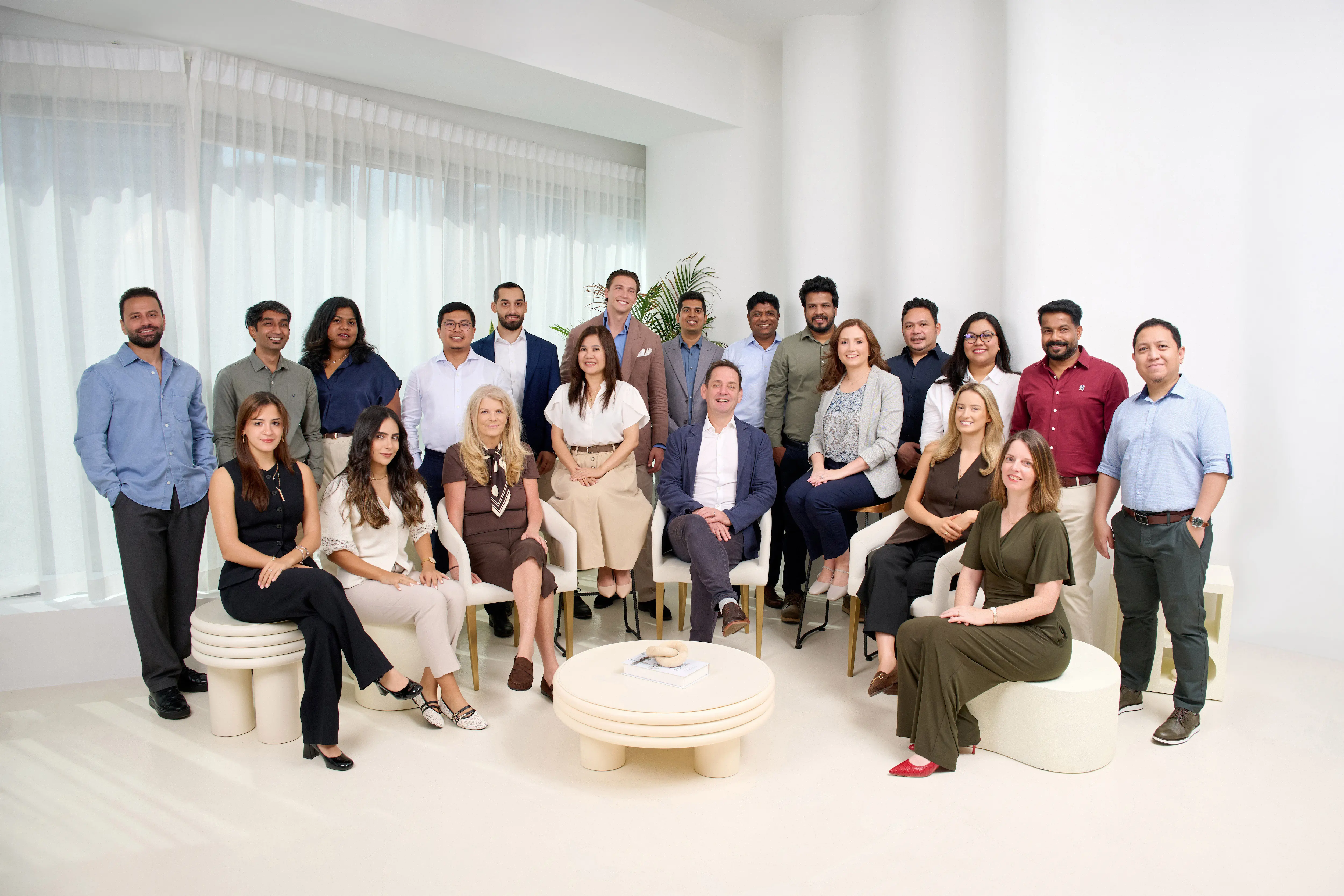 Group of twenty diverse professionals posing in a bright, modern room with white walls and sheer curtains, some standing and others seated on modern, round stools and chairs around a low, round table with a book and decorative item on it.