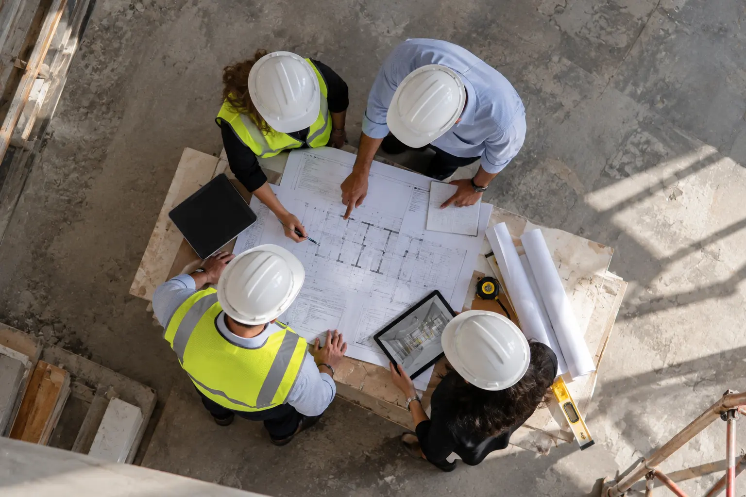 Four construction professionals wearing white hard hats gathered around a table reviewing blueprints and plans, with one person holding a tablet displaying a building model, on a concrete floor at a construction site.
