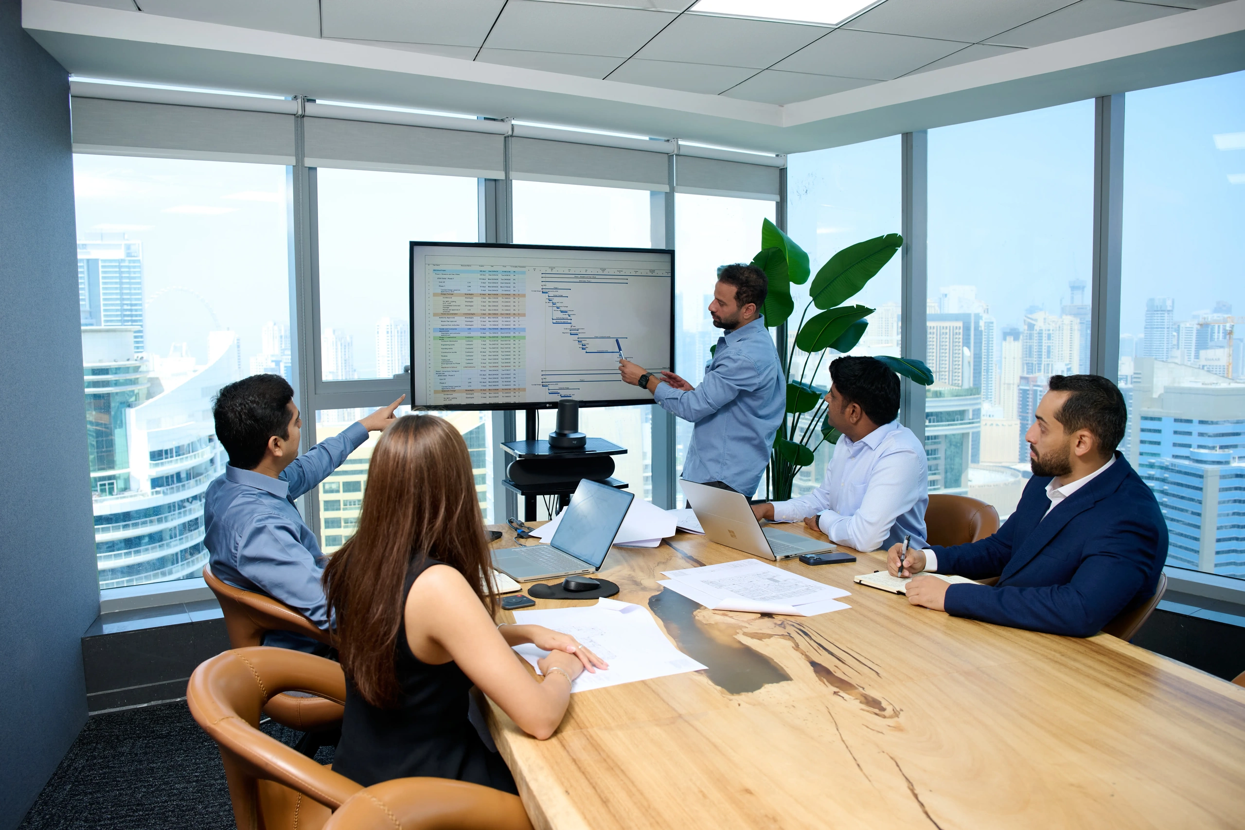 Four business professionals in a modern office with large windows overlooking a cityscape attend a presentation where a man points at a screen displaying a project timeline chart and spreadsheet data.