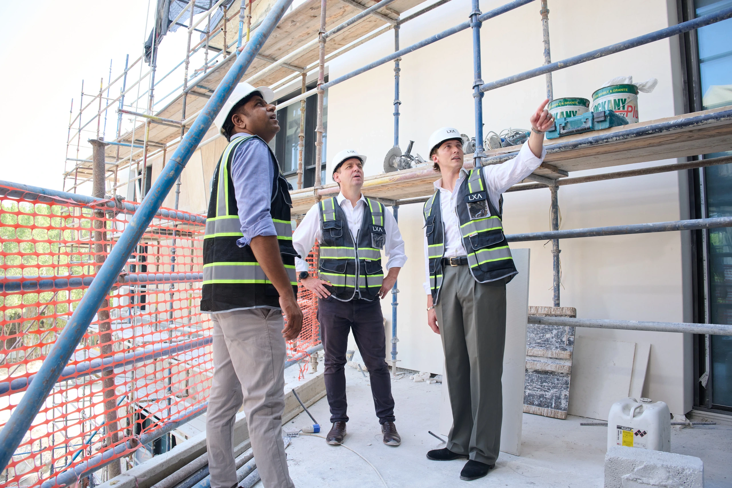 Three construction workers wearing white hard hats and reflective vests stand on a scaffolded building site, one pointing upwards while the others look in the same direction.