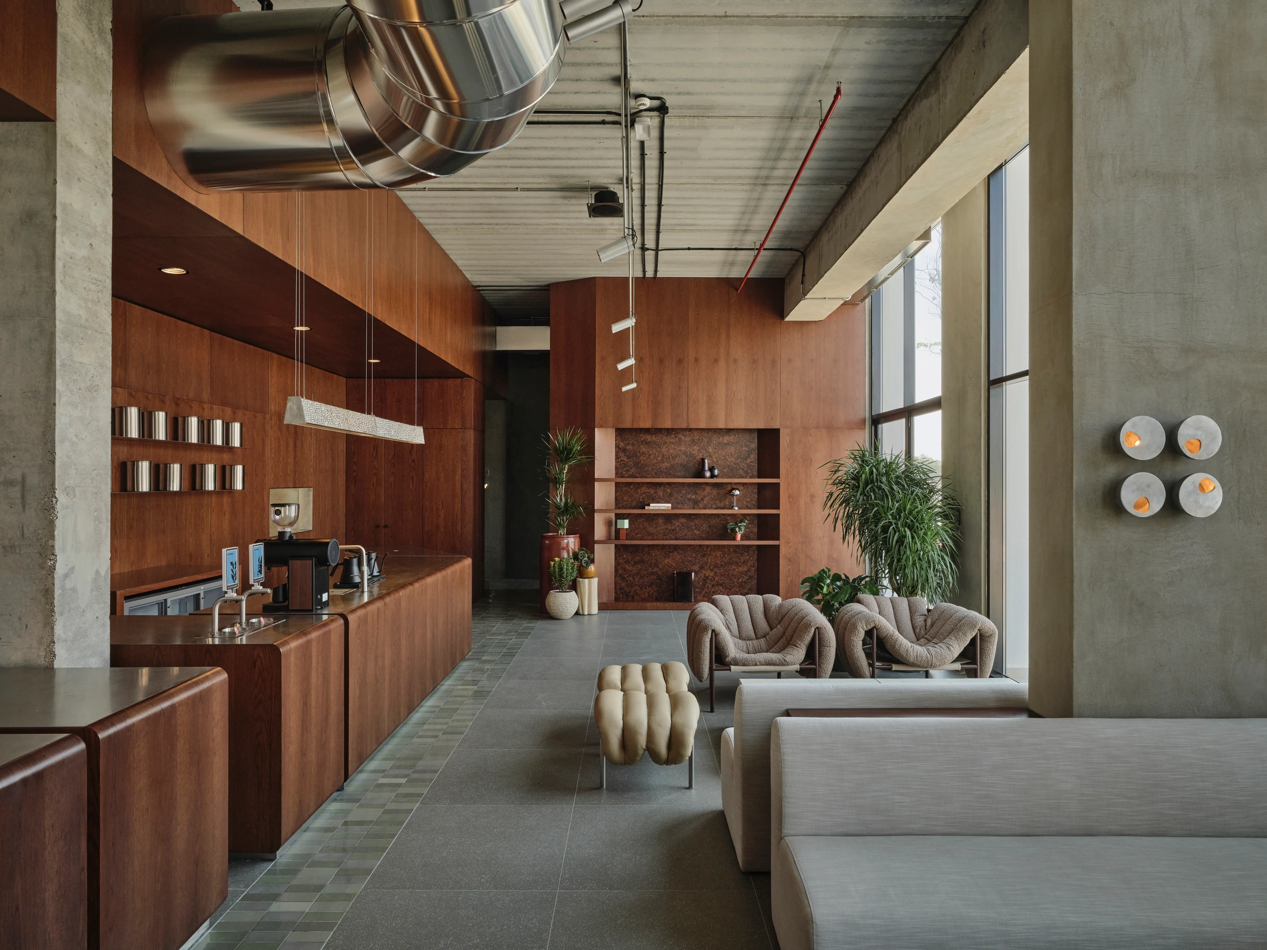 Modern interior space with a wooden counter on the left featuring a coffee machine, two beige cushioned armchairs and a unique padded bench in the center, large gray sofas on the right, tall windows with natural light, green plants near the back wooden wall with shelves, and exposed concrete ceiling with metallic ductwork.