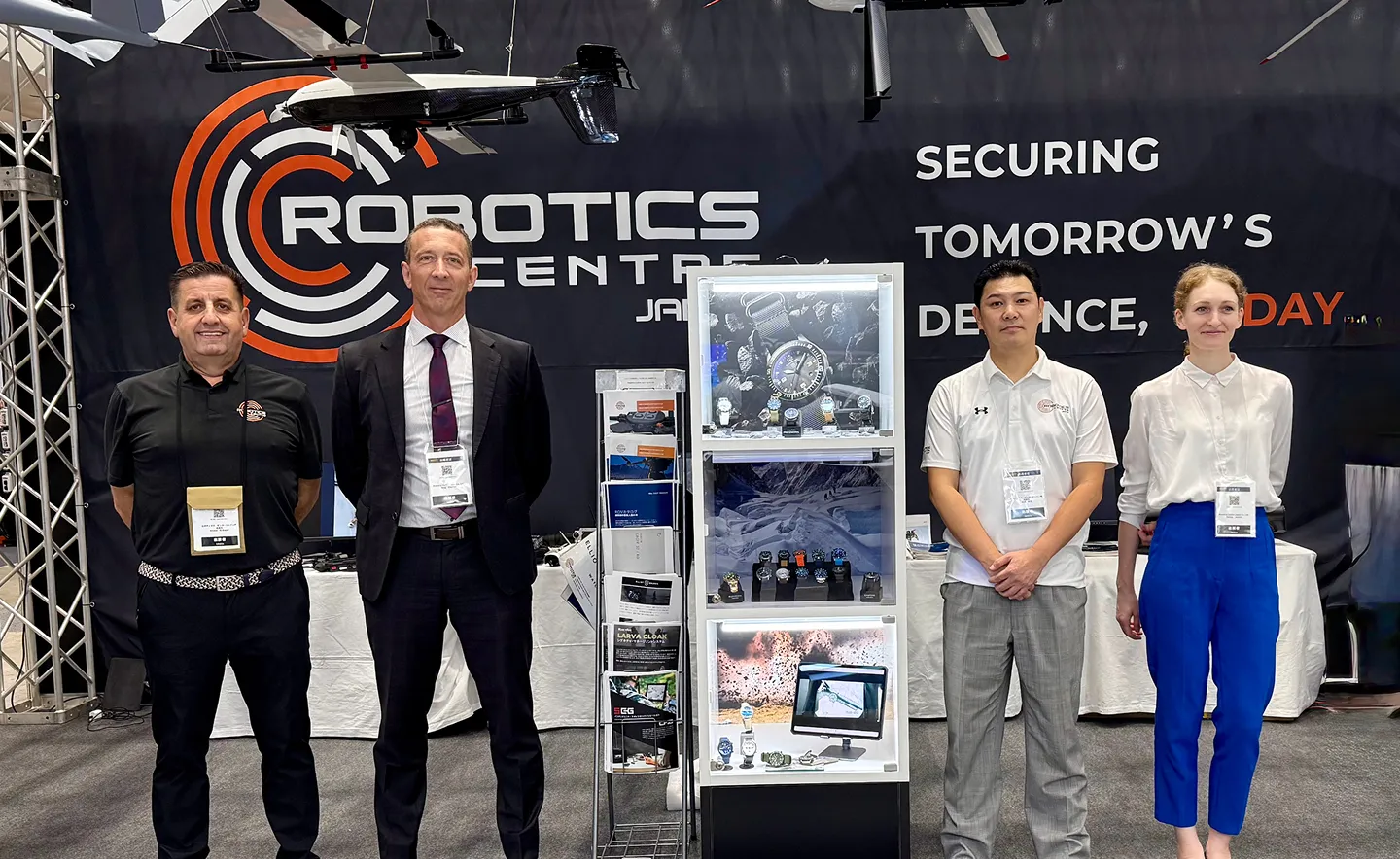4人の男女がロボティクスセンターの展示ブースの前に立っている。黒い背景には「SECURING TOMORROW'S DEFENCE, TODAY」と書かれており、ロボットのドローンが上部に展示されている。中央には時計の展示ケースが照明付きで置かれている。