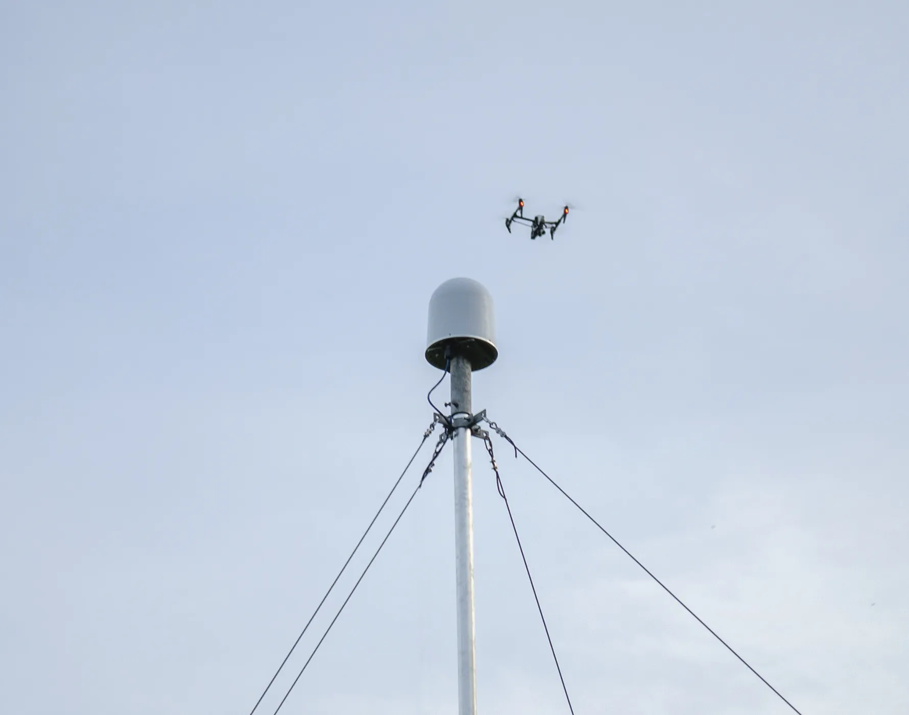 空に浮かぶ赤いライトがついた無人ドローンと、それを取り巻くワイヤーで支えられた白い円筒形のアンテナがある金属ポール。