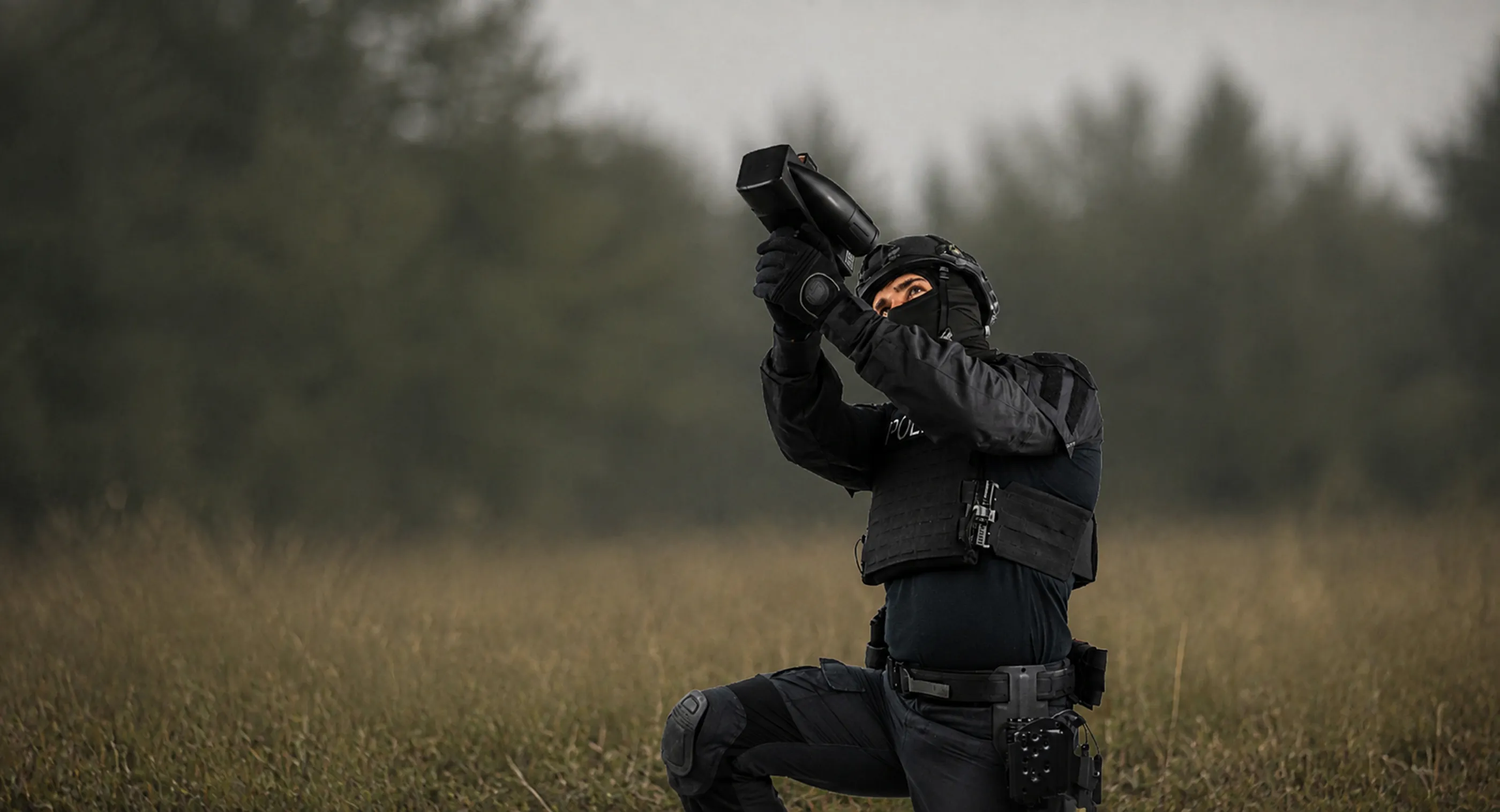 黒い戦術用装備とヘルメットを身に着けた警察官が、草原にひざまずきながら黒いデバイスを空に向けて構えている。
