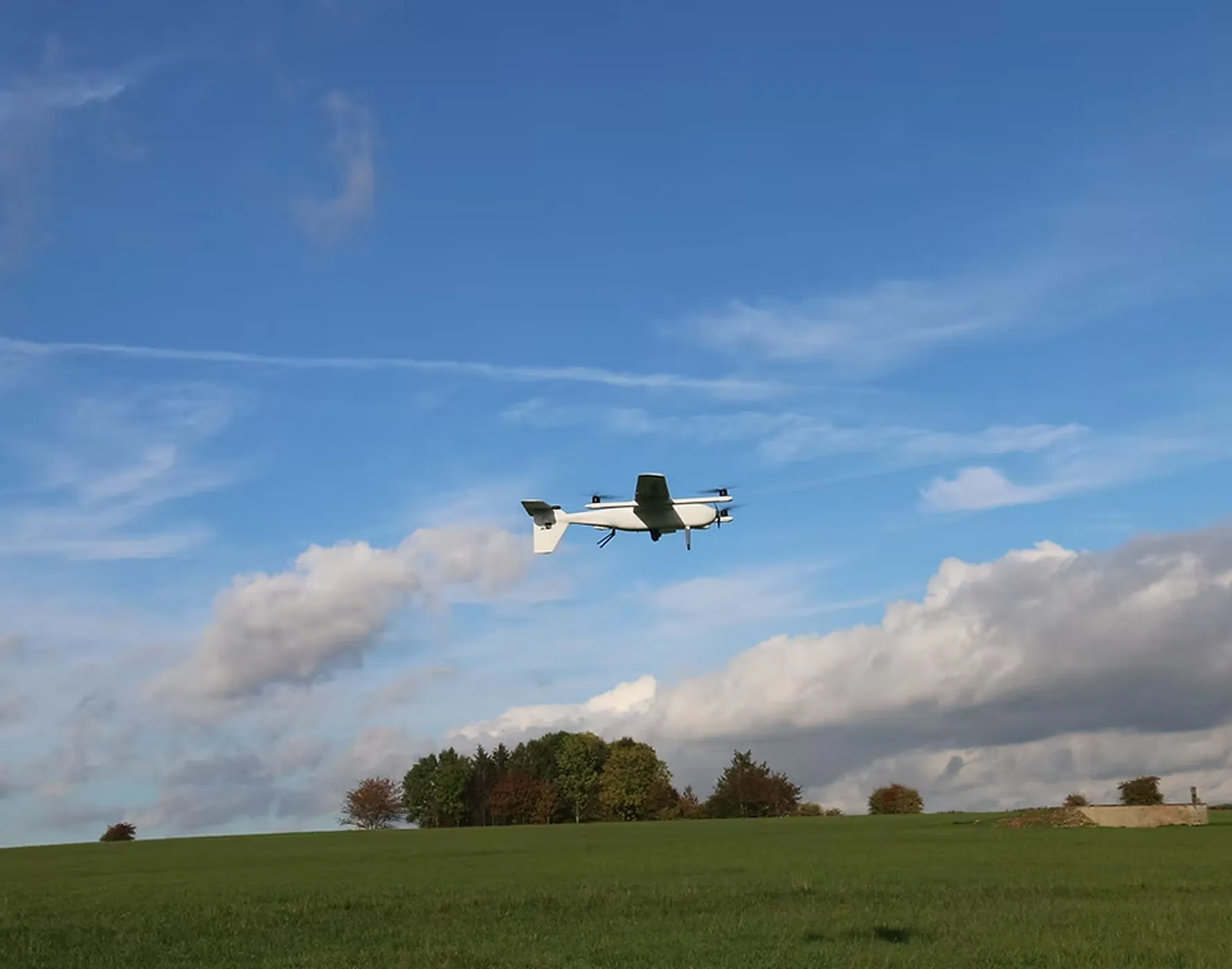 青空の下、緑の草原を背景に浮かぶ白い無人機(ドローン)。