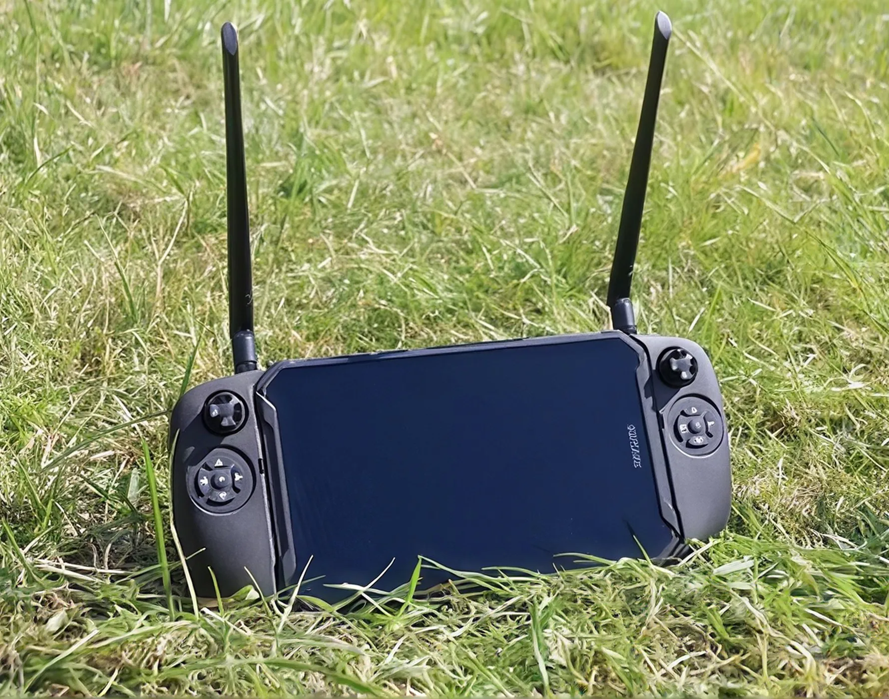 屋外の草の上に置かれた黒い携帯ゲームコントローラー、両側にスティックとボタンがあり、上部に2本のアンテナが立っている。