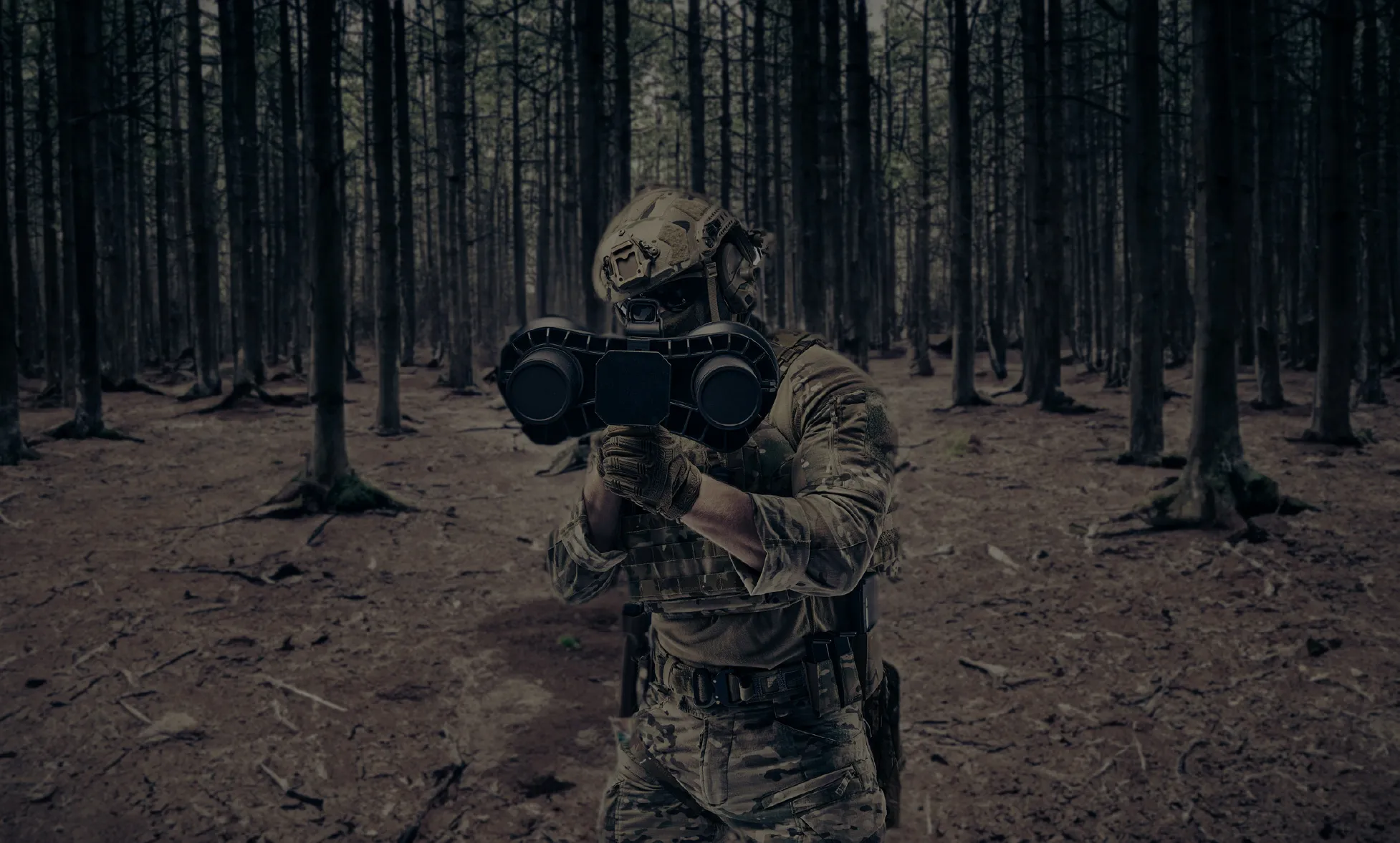 迷彩服とヘルメットを着用した兵士が、武器のような大型装置を両手で構え、森林の中で前方を見据えている。