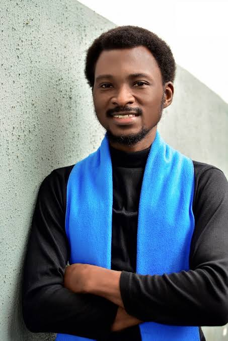 Smiling man in a black turtleneck and bright blue scarf, arms crossed, leaning against a pale textured wall.