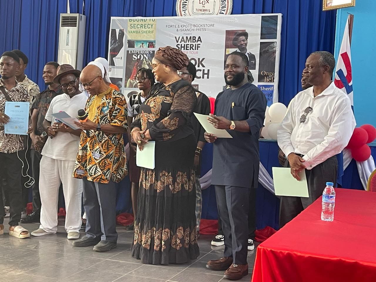 Attendees hold certificates on stage before a Vamba Sherif book launch banner from Forte & Kpoto Bookstore and Iskanchi Press.