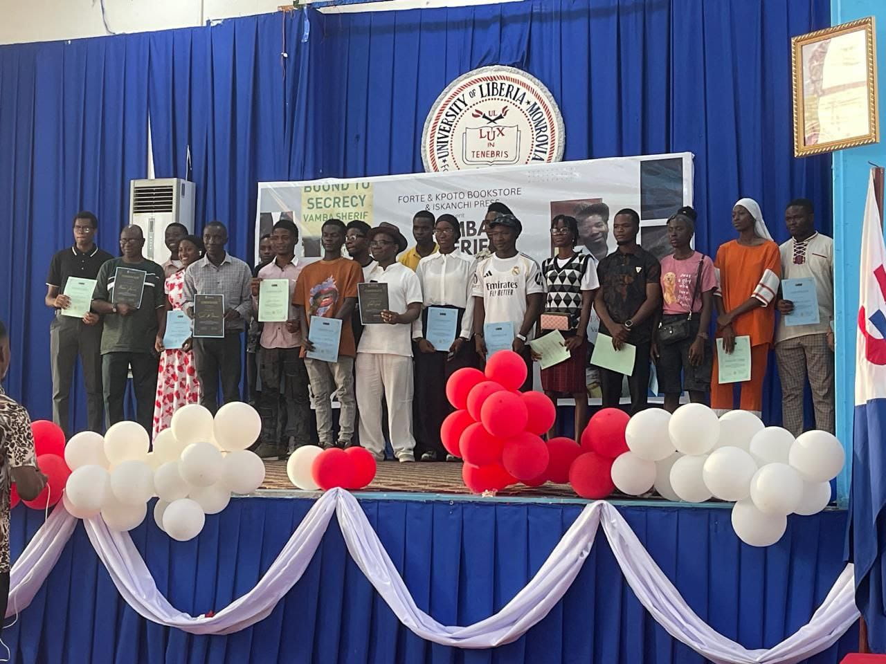 A group holding certificates on a balloon-decorated stage at the University of Liberia beneath a Vamba Sherif event banner.