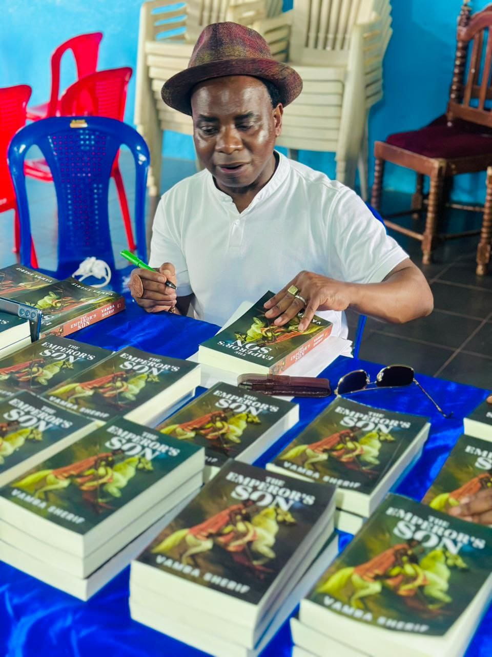 Vamba Sherif in a hat signs copies of The Emperor's Son at a blue-draped table stacked with the book.