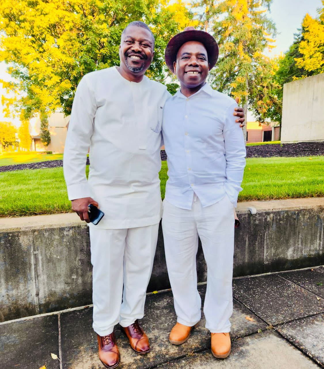 Two smiling men in white outfits, one in a brown hat, standing by a low concrete wall with autumn trees behind.