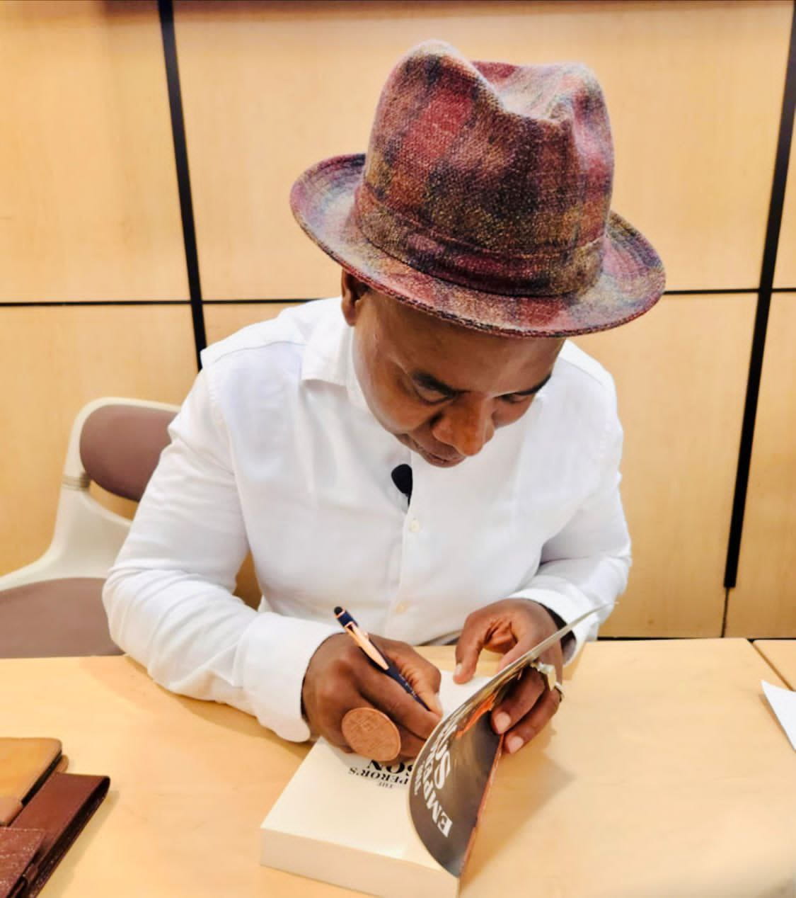 A man in a white shirt and plaid fedora signs a copy of The Emperor's Son at a wooden table in a wood-panelled room.
