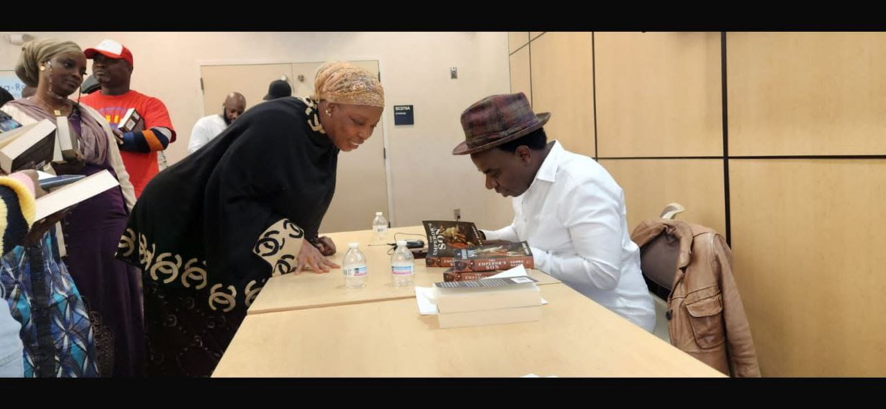 An author in a hat signs copies of The Emperor's Son at a table while a reader in a headwrap leans in and others queue behind.