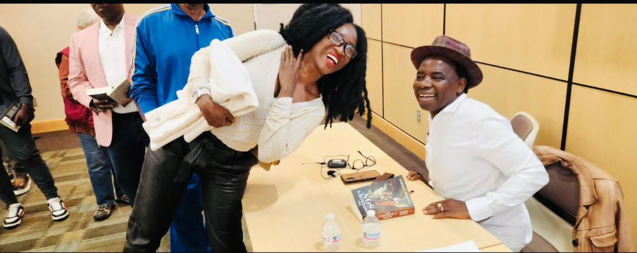 A smiling woman leans toward a seated author in a hat at a book-signing table as attendees line up with books behind her.