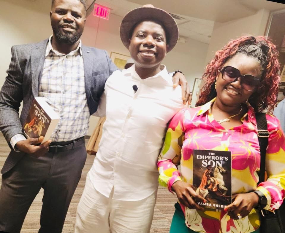 Three smiling people pose indoors, two holding copies of The Emperor's Son by Vamba Sherif, one wearing a hat.