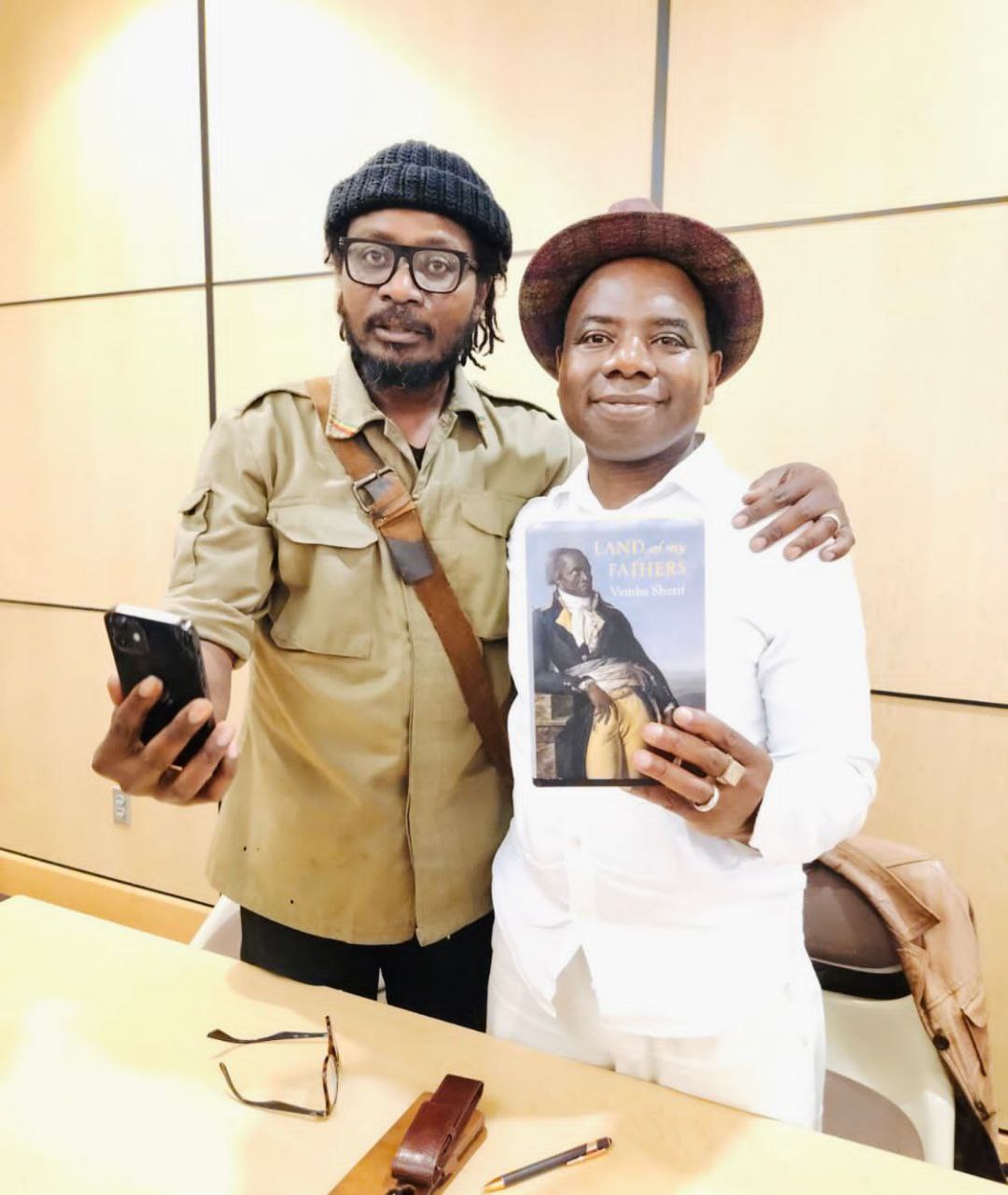 Two men pose indoors by a table, one in a hat and white shirt holding a copy of Land of My Fathers by Vamba Sherif.