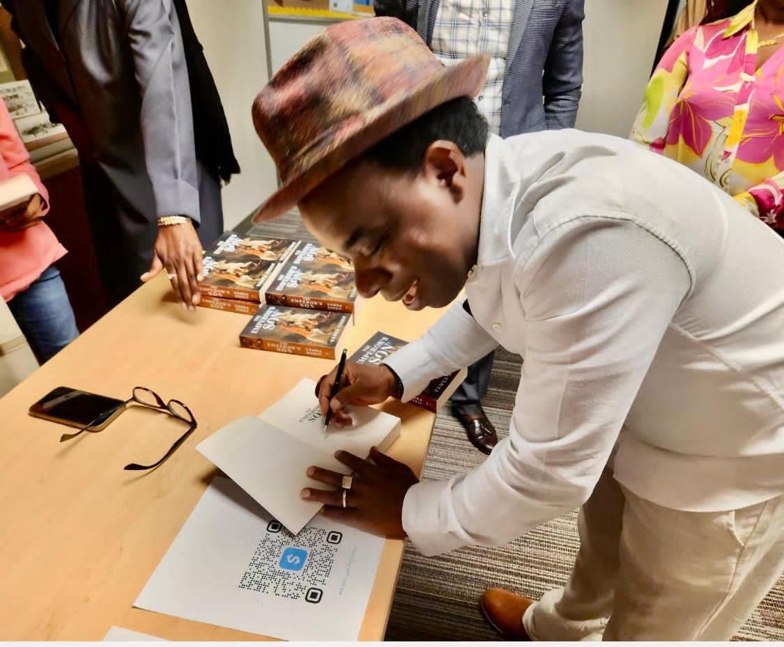 A person in a patterned hat and white shirt signs a copy of The Emperor's Son at a table stacked with the book.