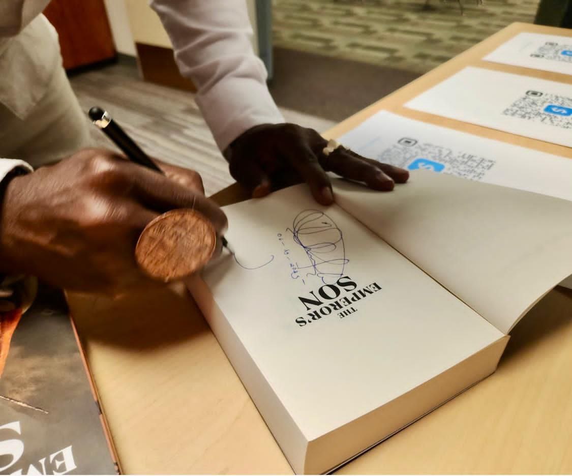 Hands signing the title page of The Emperor's Son with a pen, beside printed QR code cards on a wooden table.