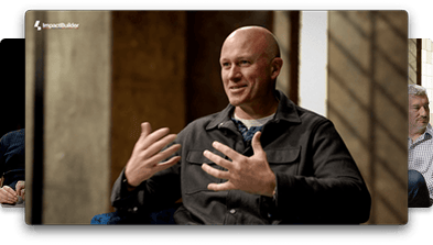 A bald man wearing a dark jacket gestures with his hands while speaking in an indoor setting with another person partially visible in the background.