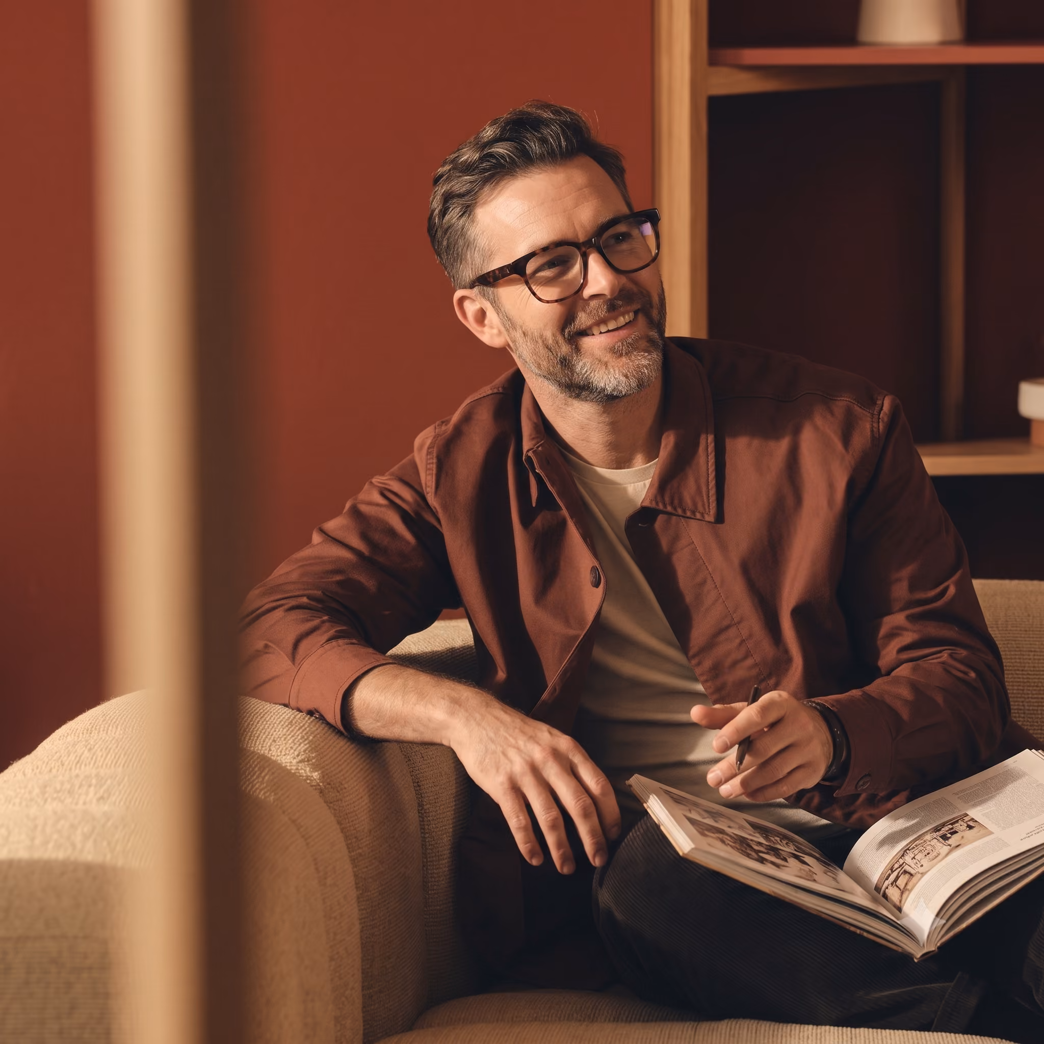Smiling man with glasses sitting on a couch reading a book in a warm-toned room.