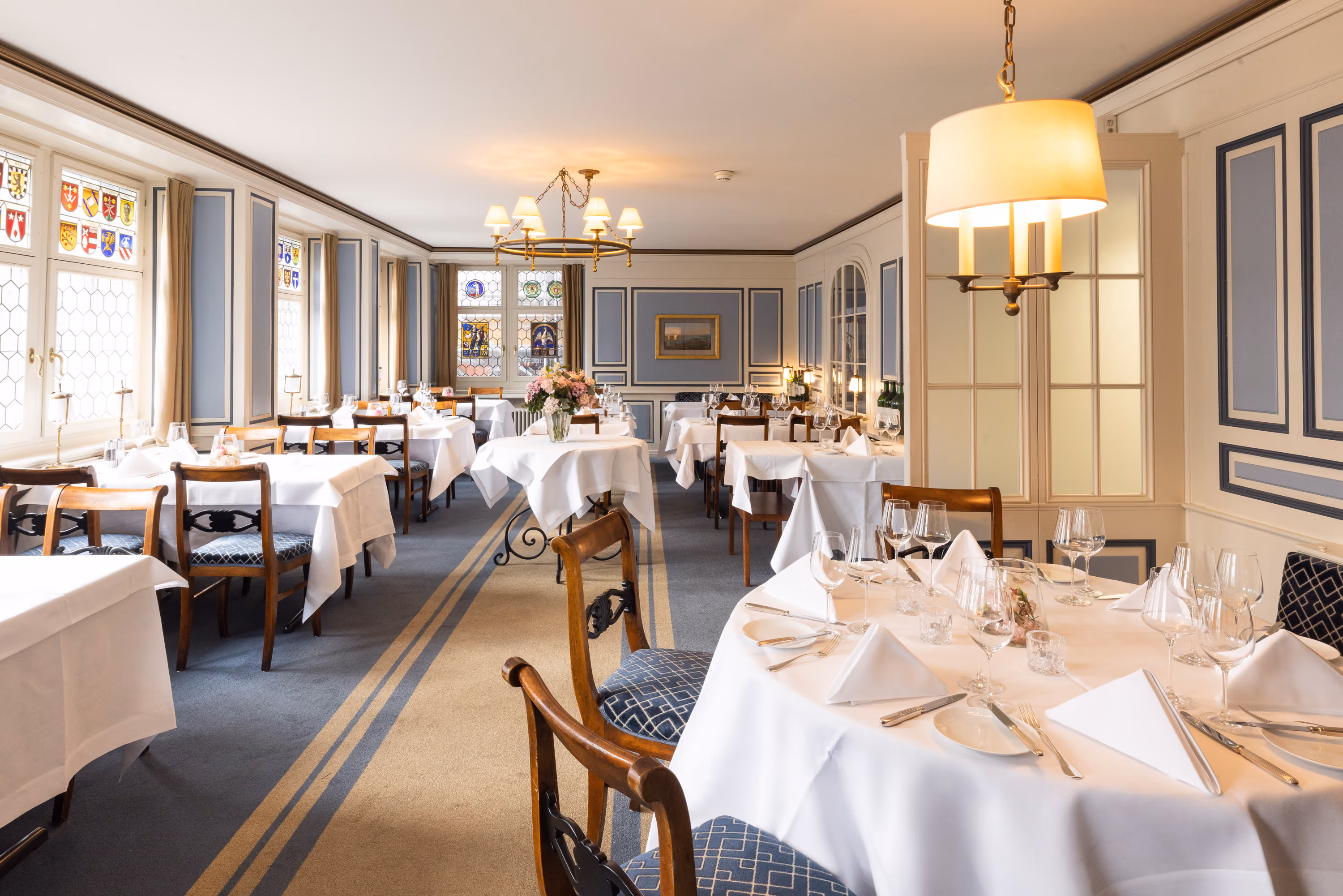 Heller Restaurantraum im Zunfthaus zur Waag mit weiss gedeckten Tischen, Kronleuchtern und Wappenscheiben