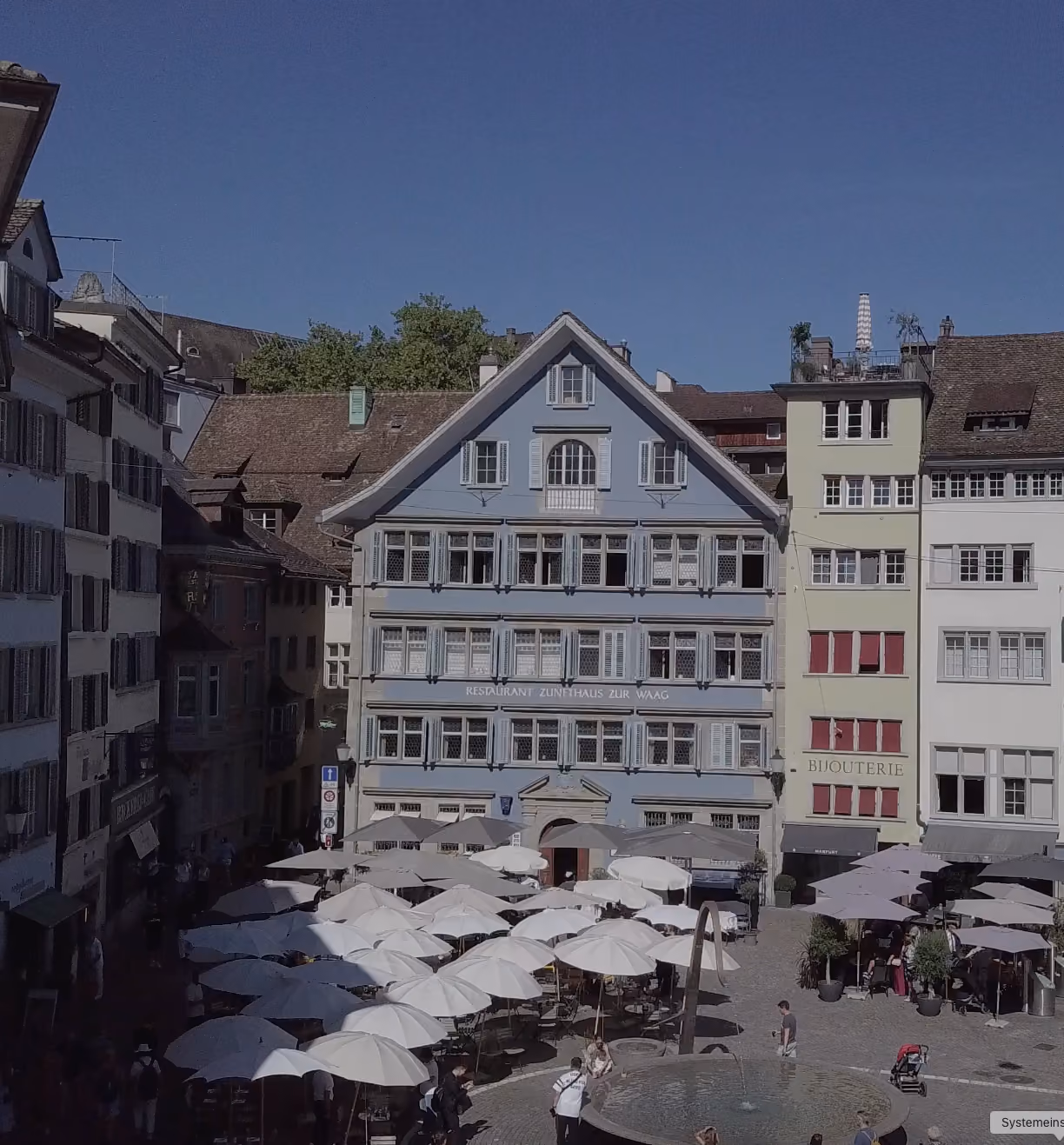 Hellblaue Fassade des Zunfthauses zur Waag am Münsterhof mit Brunnen und Sonnenschirmen der Terrasse