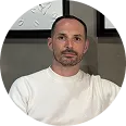 Portrait of a man with short dark hair and a trimmed beard wearing a white sweater, posing indoors against a wall with a clock partially visible behind him.