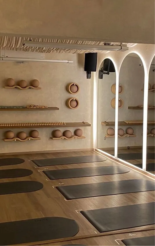 A yoga or Pilates studio with wooden floors, black exercise mats arranged in rows, and beige exercise balls on wooden shelves along the wall; large arched mirrors with backlighting reflect the room.
