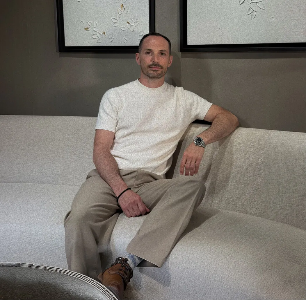 Man with short dark hair and trimmed beard wearing a white short-sleeve sweater, beige pants, brown shoes, and a watch on his left wrist, sitting on a beige couch against a muted wall with framed artwork.