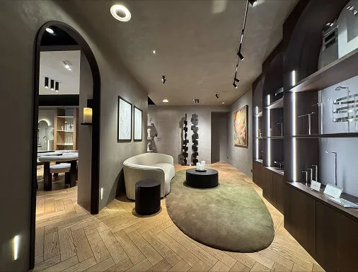 Modern interior room with wood herringbone floor, beige curved sofa, round black coffee table, green oval rug, and backlit shelves displaying faucets and fixtures.