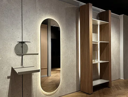 Modern interior wall featuring an oval mirror with backlighting, a minimalist white floating shelf, and a tall wooden shelving unit with glass shelves and a few clear glass vases inside.