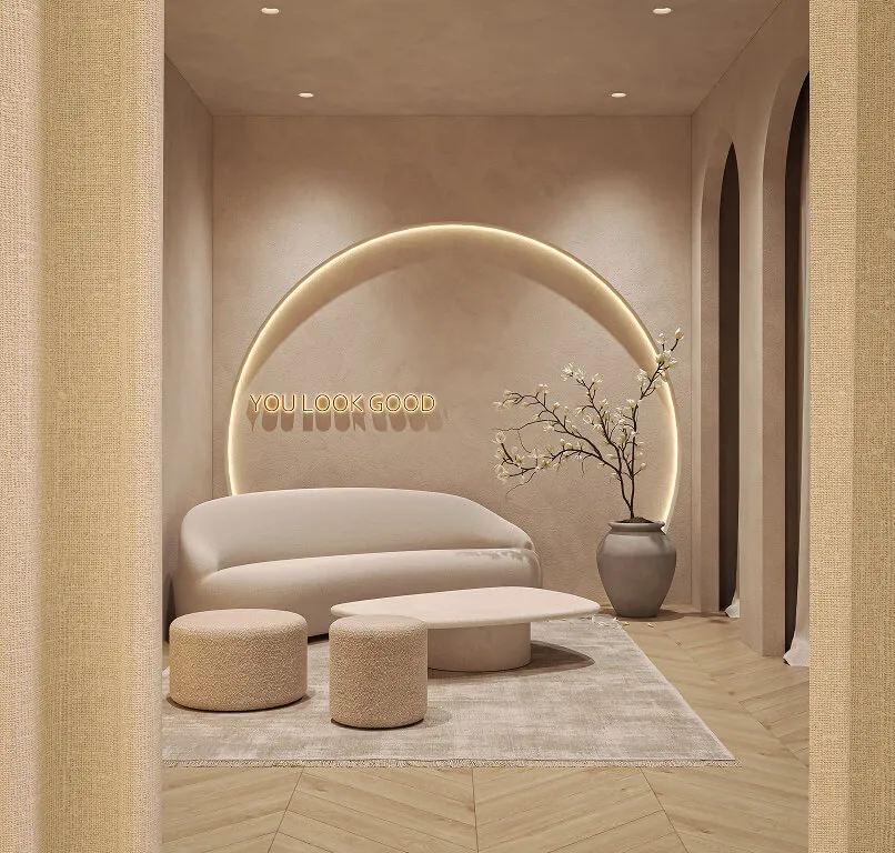 Minimalist beige lounge area with a curved sofa, two round ottomans, and a rounded rectangular coffee table on a light rug, set against a wall with a glowing semicircle light and text reading 'YOU LOOK GOOD'. A vase with branches and flowers is placed near three arch openings on the right.