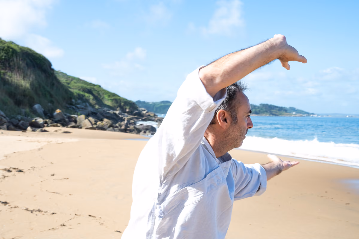 Un homme en chemise blanche pratique un mouvement de baguazhang sur une plage sablonneuse avec des collines verdoyantes en arrière-plan et l'océan sous un ciel bleu.