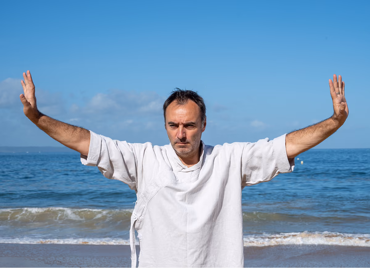 Homme pratiquant le tai-chi avec les bras levés et les paumes ouvertes sur une plage, avec la mer et le ciel bleu en arrière-plan.