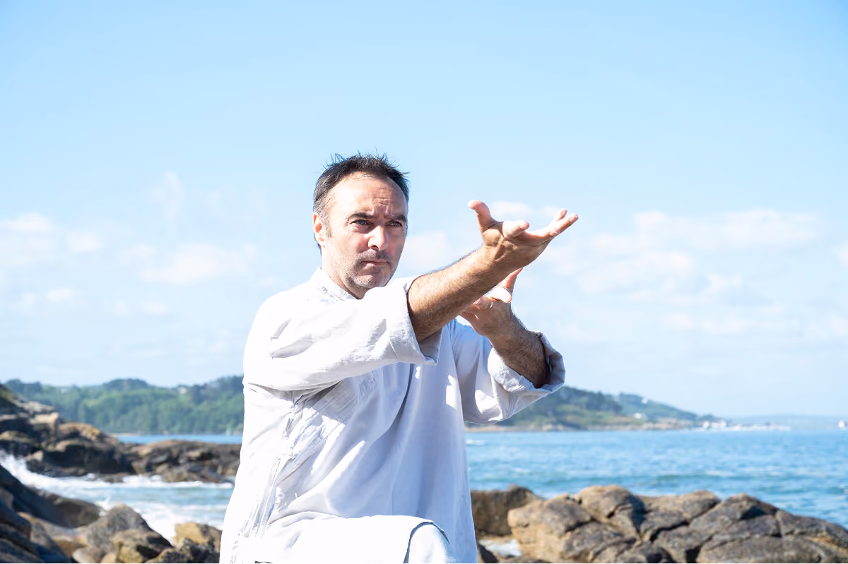 Homme en tenue blanche pratique le Qi Gong sur des rochers au bord de la mer avec un ciel bleu et des collines en arrière-plan.