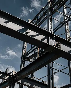 Structural steel framework under construction with intersecting beams and bolts, viewed from low angle against a partly cloudy sky