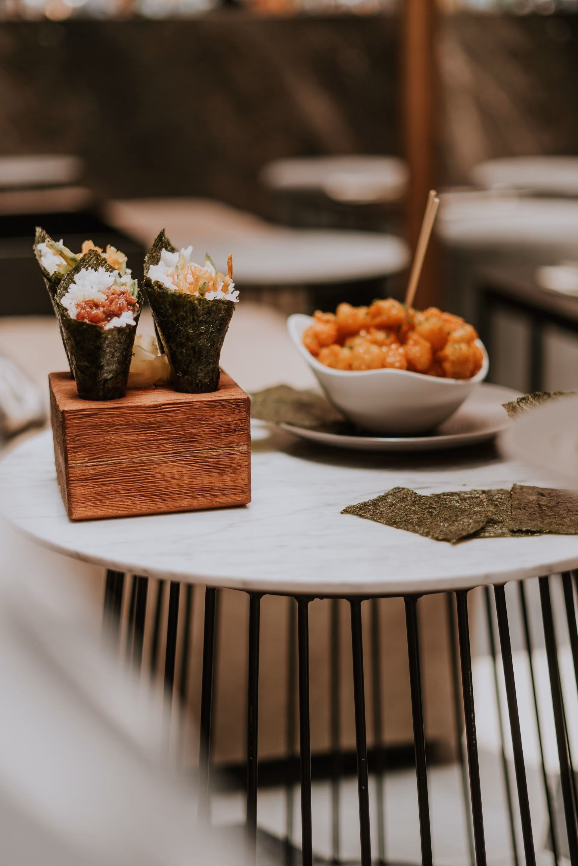 Two seaweed hand rolls filled with rice and seafood placed on a wooden block on a round white table, next to a white bowl with fried food and a skewer, with a few seaweed sheets on the table.