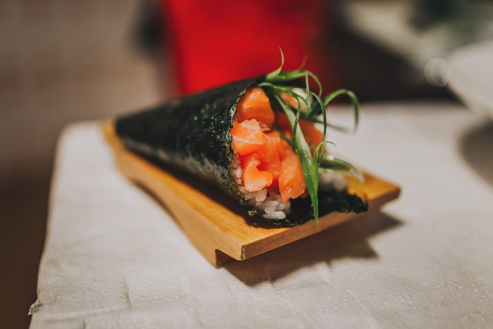Close-up of a salmon sushi hand roll with seaweed wrap, white rice, and green garnishes on a wooden serving block.