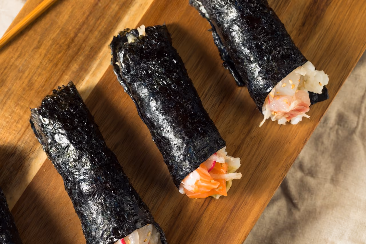 Four sushi hand rolls wrapped in dark seaweed on a wooden board, filled with rice and various ingredients like crab and salmon.