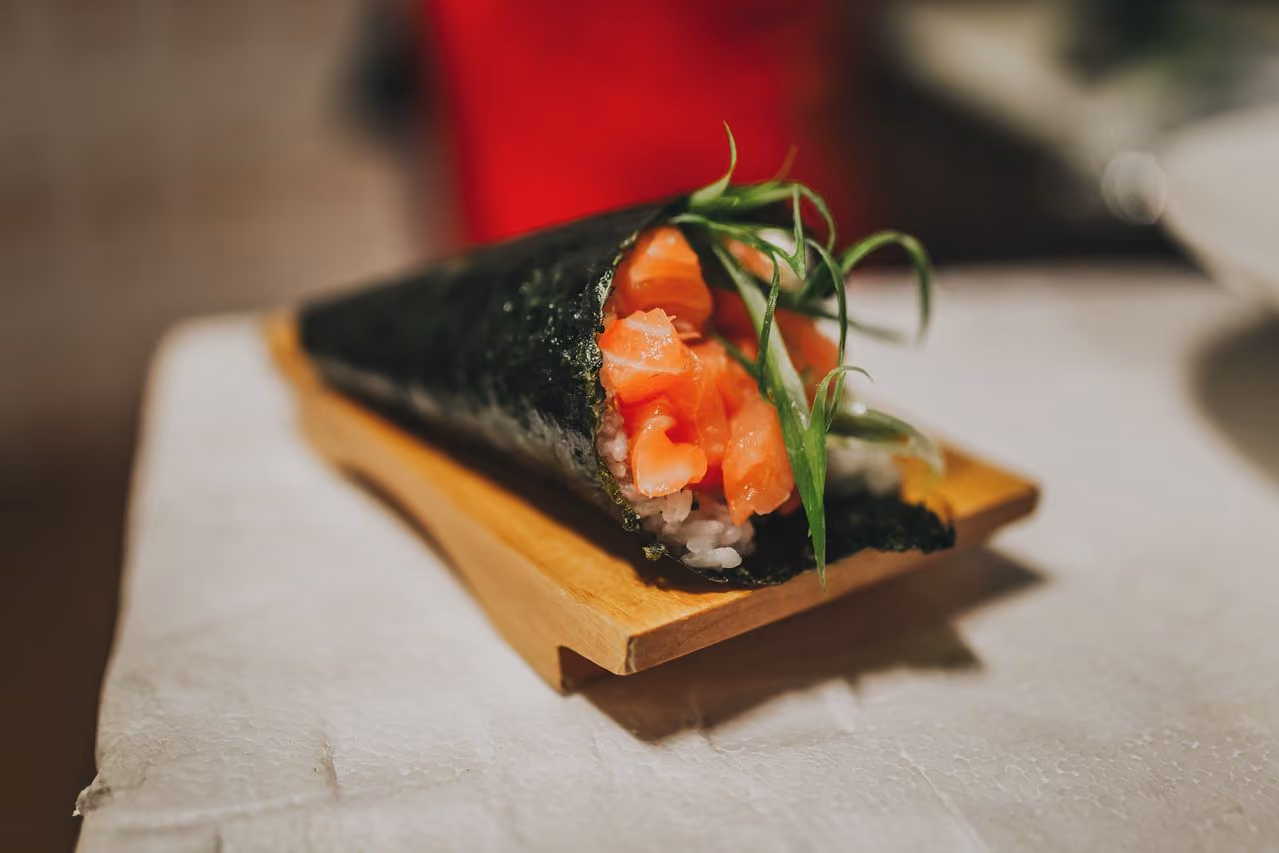 A fresh salmon temaki sushi cone wrapped in nori seaweed, filled with rice and topped with green garnish, resting on a small wooden block on a white surface.