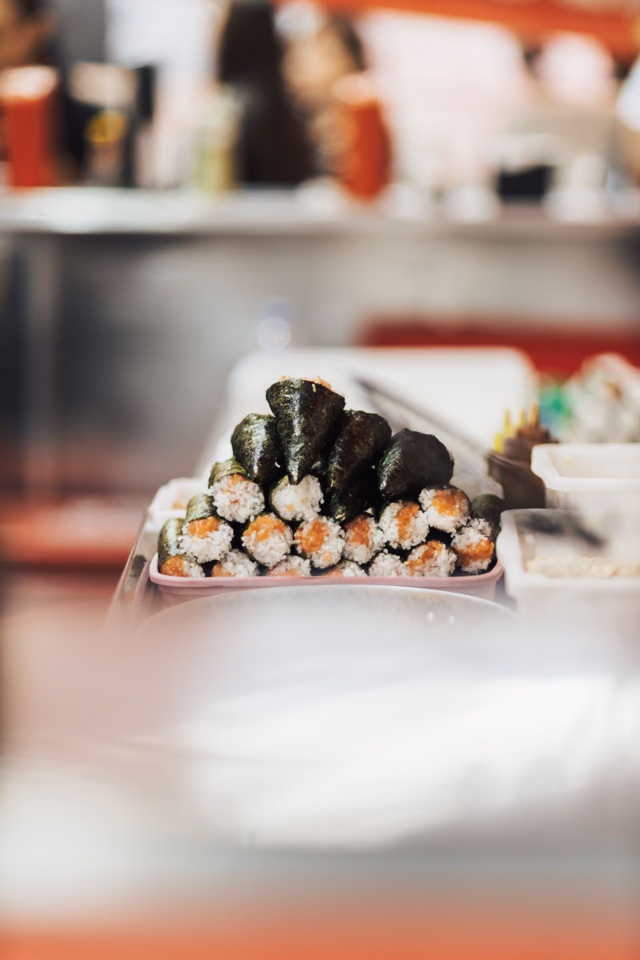 Stacked sushi rolls and hand rolls on a tray with a blurred background of a kitchen or restaurant setting.