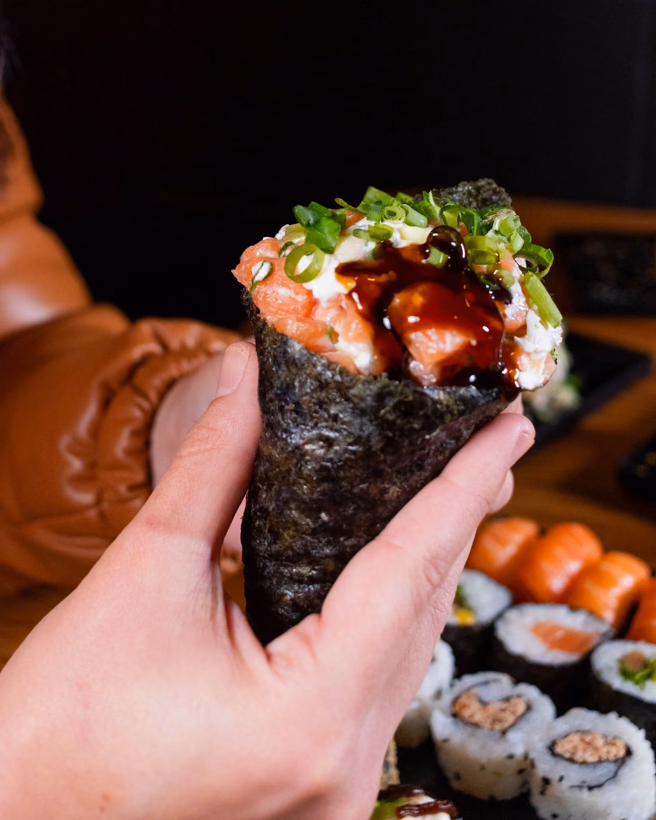 Hand holding a sushi hand roll wrapped in seaweed, filled with salmon, mayonnaise, green onions, and drizzled with dark sauce, with various sushi rolls blurred in the background.