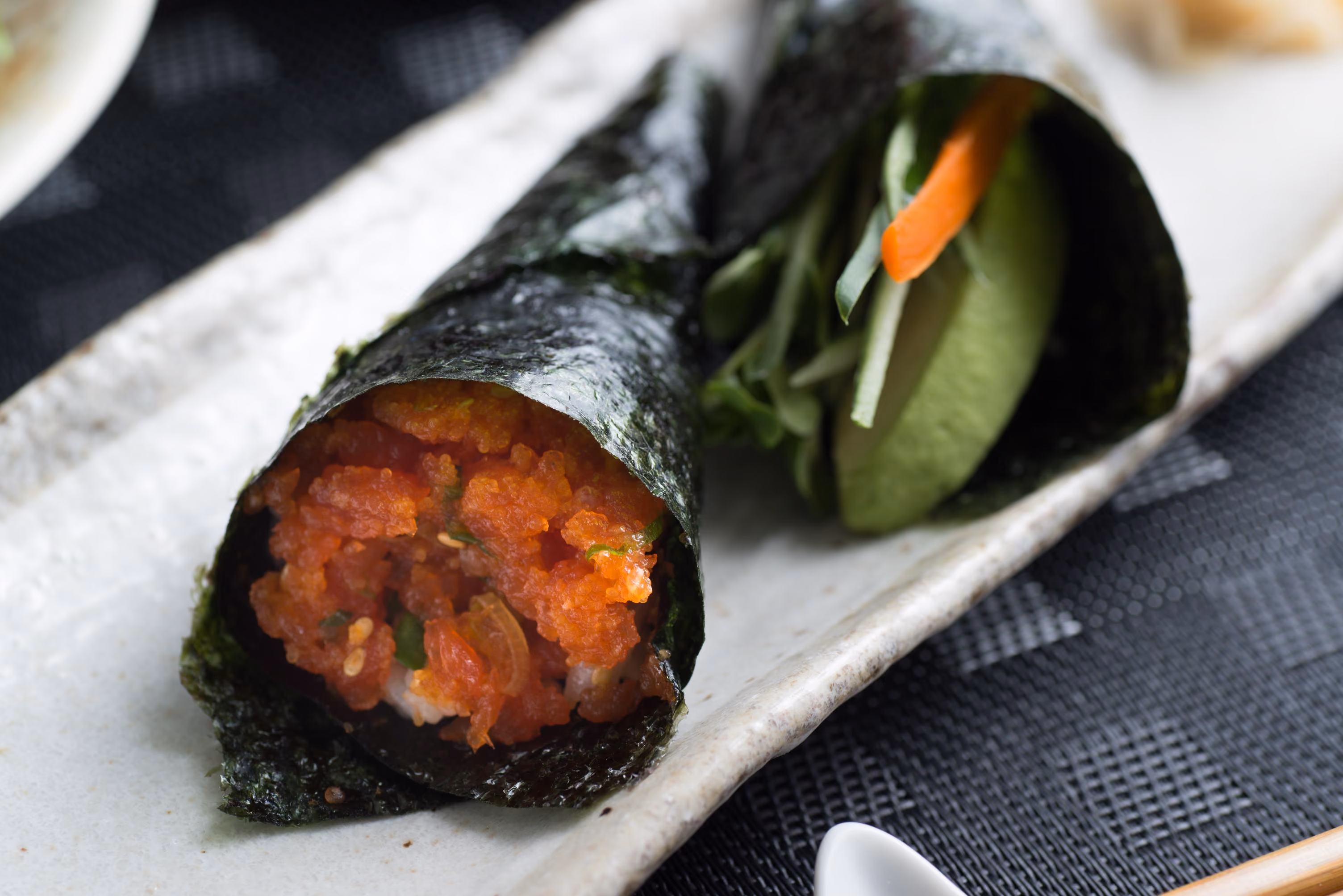 Two sushi hand rolls on a rectangular ceramic plate; one contains spicy minced tuna, the other has sliced avocado, cucumber, carrot, and leafy greens wrapped in seaweed.