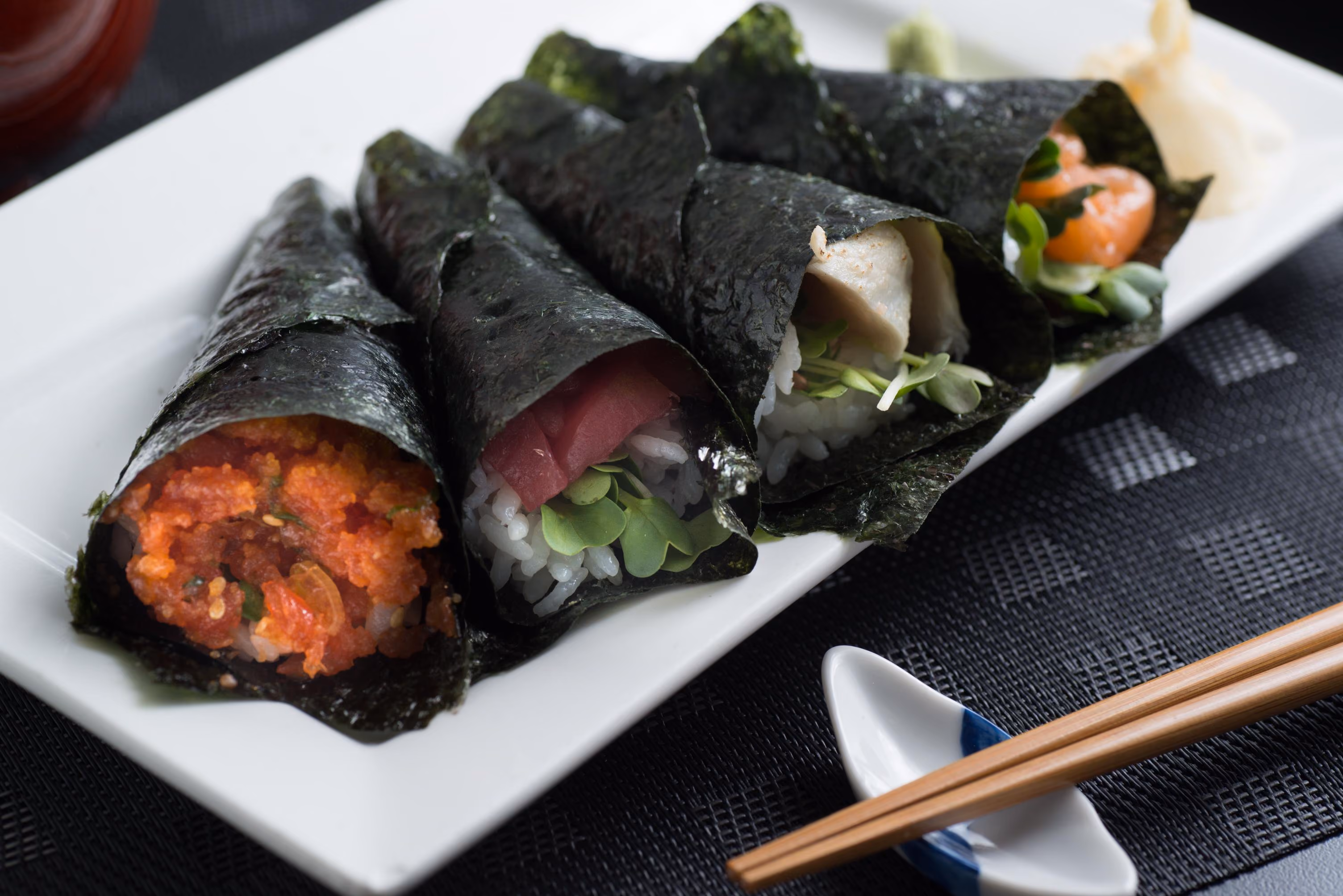 Four hand rolls (temaki sushi) on a white rectangular plate, each wrapped in seaweed and filled with rice and assorted ingredients including spicy tuna, raw tuna, cooked fish, and salmon with green garnish, with wooden chopsticks resting on a holder beside the plate.
