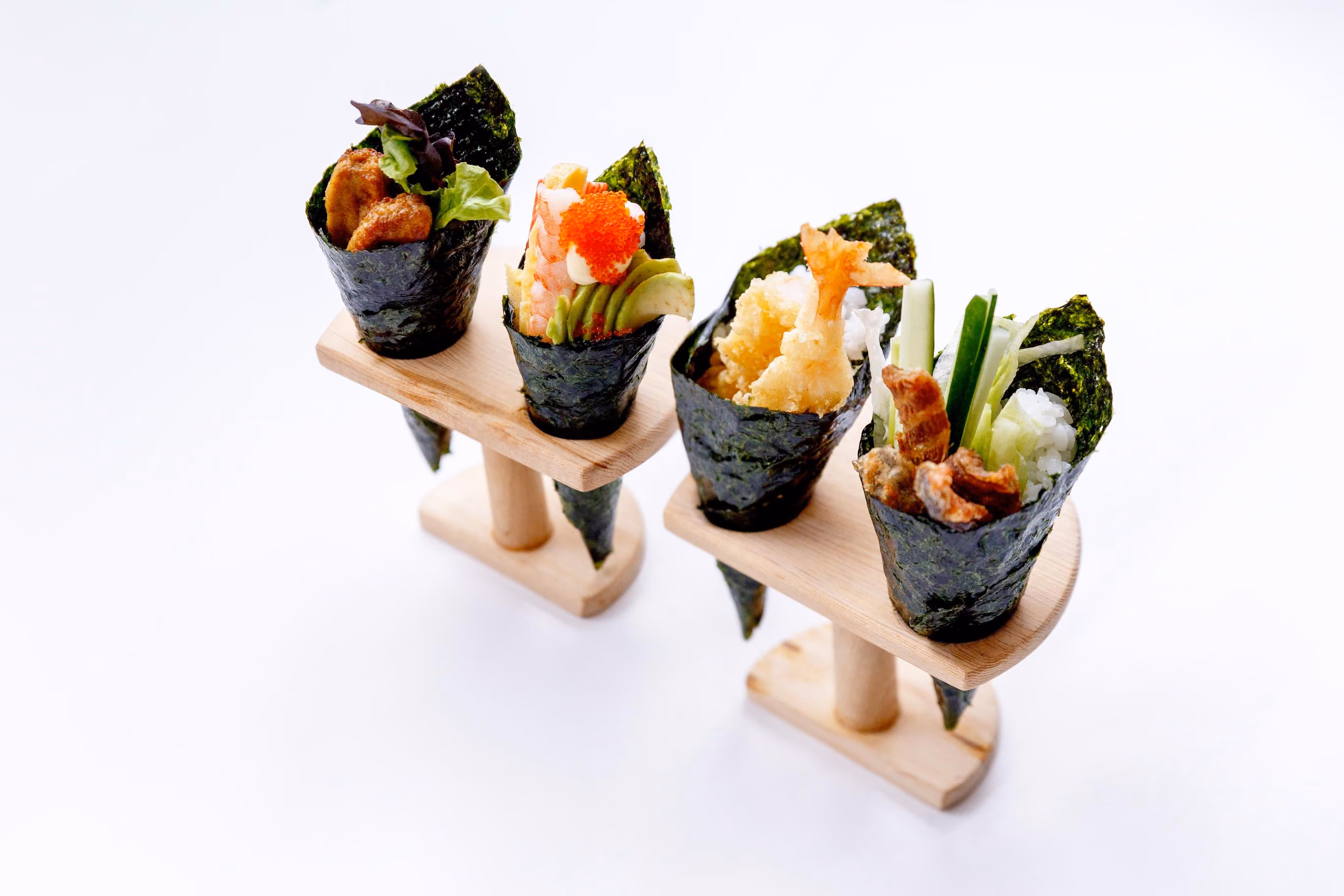 Four assorted hand rolls sushi cones with seaweed wrap, filled with ingredients including tempura shrimp, avocado, fish roe, fried chicken, and vegetables, displayed upright on two wooden stands against a white background.