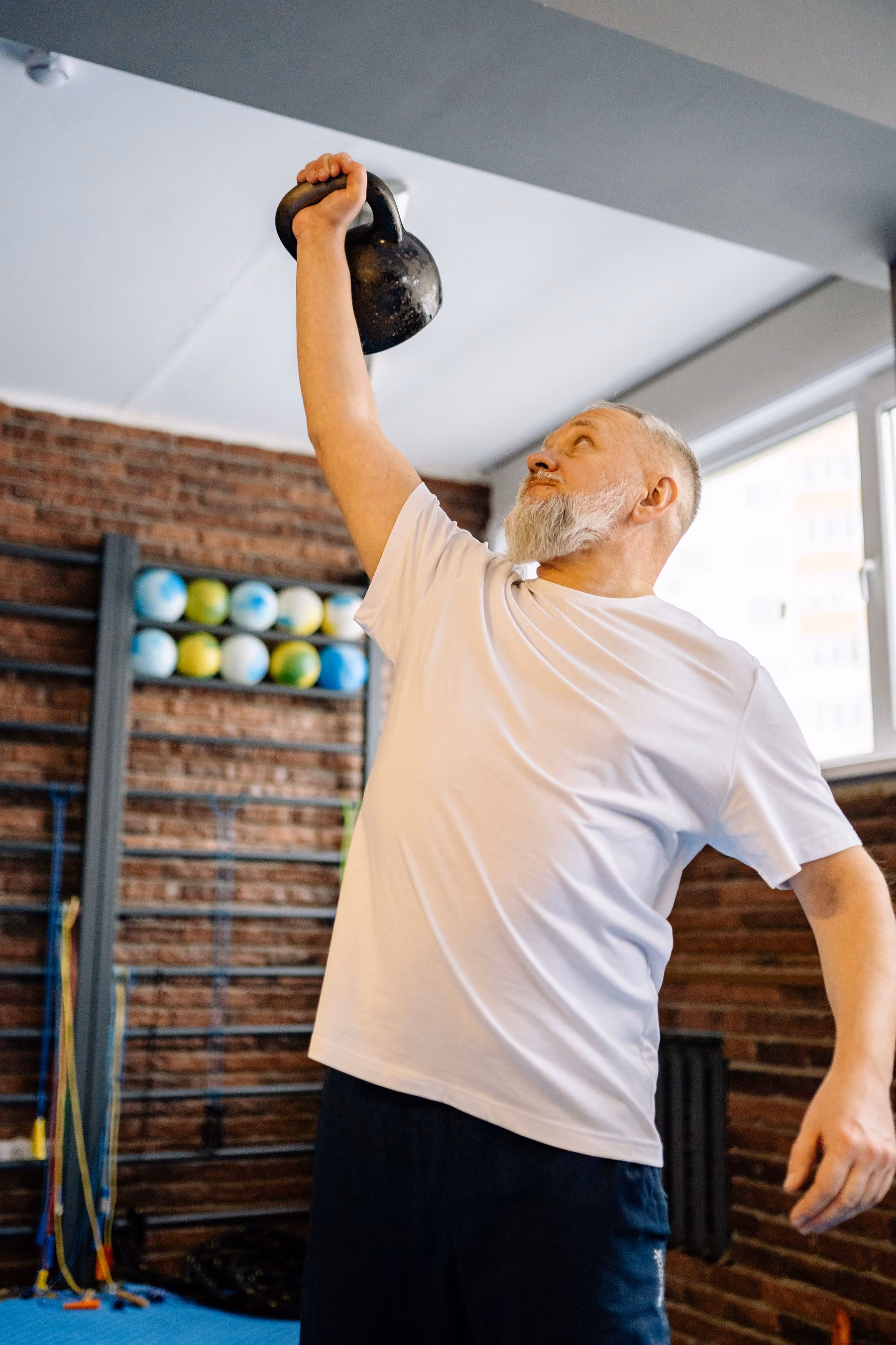 An older man with a white beard wearing a white t-shirt lifting a black kettlebell overhead in a gym with brick walls and exercise balls in the background.