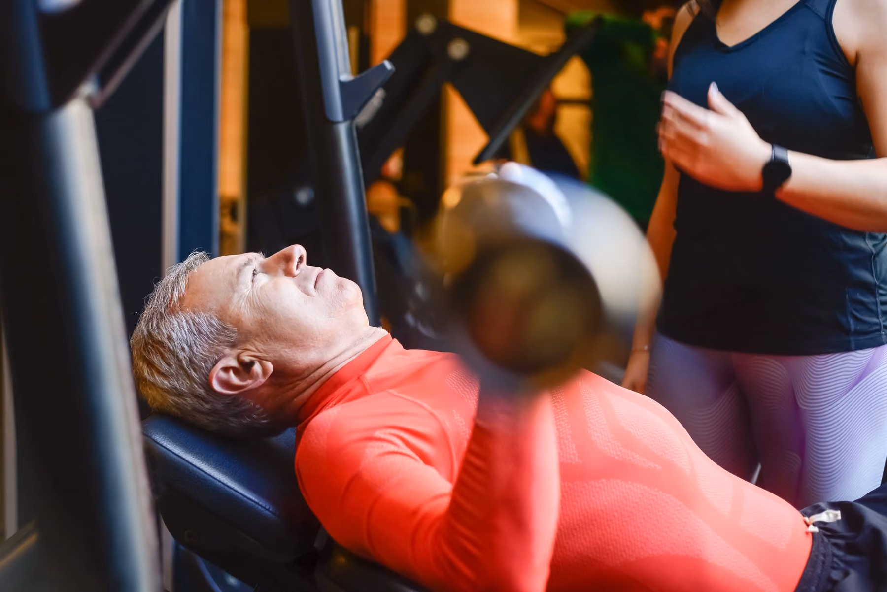 Older man in a red long-sleeve shirt lifting a dumbbell while lying on an inclined bench at the gym, with a trainer standing nearby.